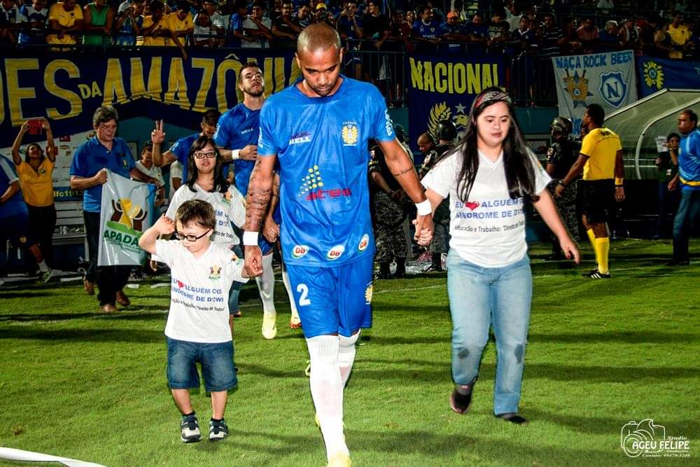 Jogador de futebol do time Nacional entrando em campo de mãos dadas com uma criança e uma adolescente Down, vestidos com a blusa da APADAM.